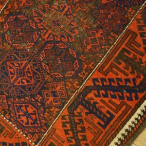Geometric woven rug corner in rust red, dark blue, and brown, lying on light wood flooring.