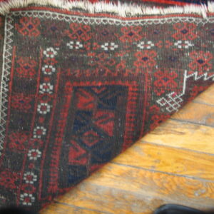 A dark red and brown geometric rug with white fringe laid diagonally on shiny hardwood floors.