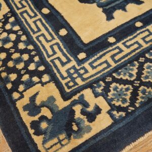 Navy and tan rug displaying geometric borders and stylized floral motifs rests on worn light wood flooring.