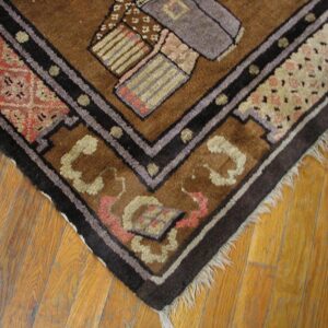 Corner of a brown geometric rug with dark borders and fringe rests on golden wood plank flooring.