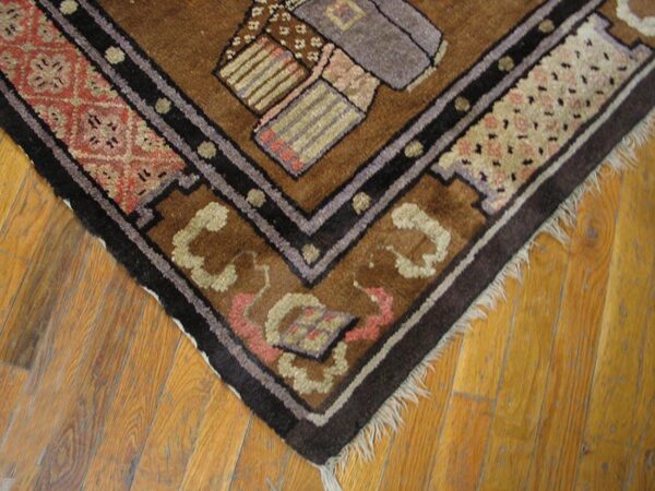 Corner of a brown geometric rug with dark borders and fringe rests on golden wood plank flooring.