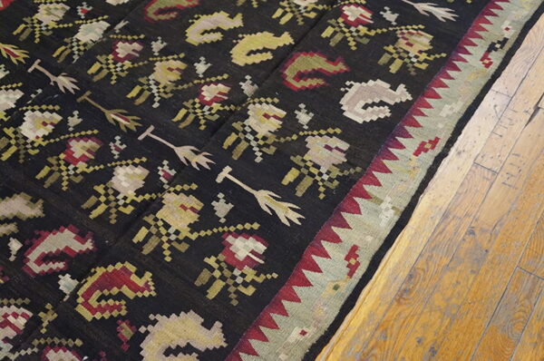 Flat-woven rug with geometric red, cream, and olive patterns on a black field and a plank wood floor.