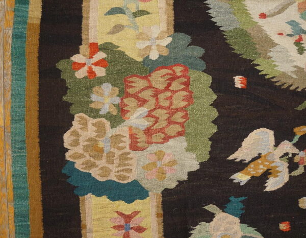 Dark flat-weave rug featuring large, colorful floral and bird patterns, resting on light wood flooring.
