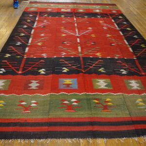 Geometric flat-weave rug featuring a red field, black stripes, green border, and stylized repeating designs on wood flooring.