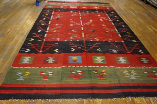 Geometric flat-weave rug featuring a red field, black stripes, green border, and stylized repeating designs on wood flooring.