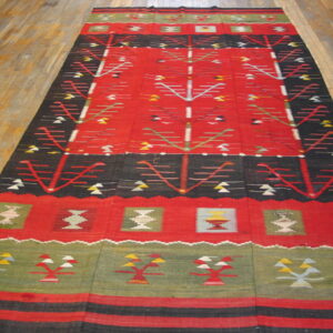 Red, black, and green flat-weave rug with stylized geometric motifs laid on light brown plank flooring.