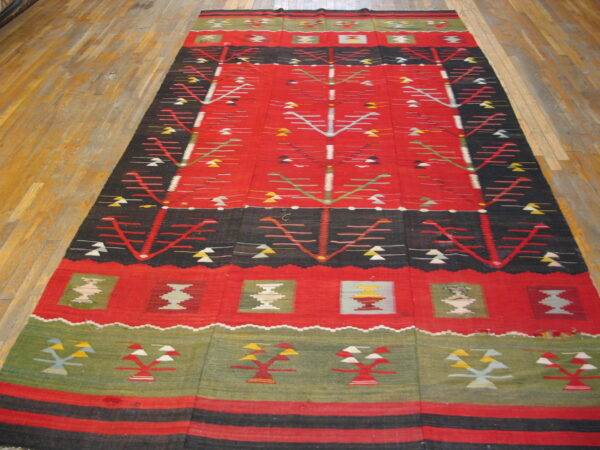Red, black, and green flat-weave rug with stylized geometric motifs laid on light brown plank flooring.