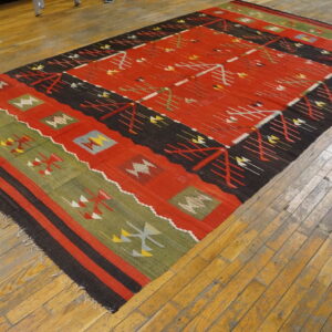Red, black, and green flat-woven rug with geometric motifs laid on rustic wooden plank flooring.