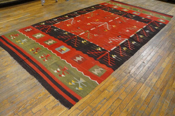 Red, black, and green flat-woven rug with geometric motifs laid on rustic wooden plank flooring.