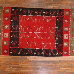 Flat-woven rug in red, black, and olive features complex geometric patterns on a distressed wooden floor.