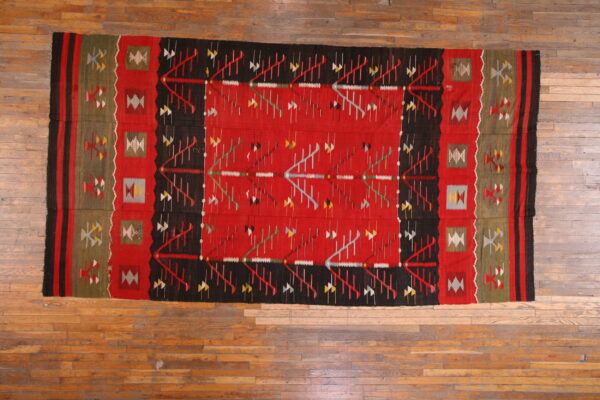 Flat-woven rug in red, black, and olive features complex geometric patterns on a distressed wooden floor.
