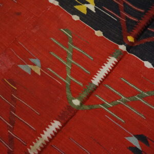 Flatweave rug close-up featuring stylized green, white, and red motifs on a deep red and dark navy background.