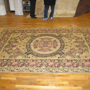 Pale gold rug with a large black and rose floral medallion displayed on a shiny wood floor.