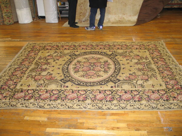 Pale gold rug with a large black and rose floral medallion displayed on a shiny wood floor.