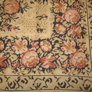 Beige rug corner with pink and red floral motifs and dark blocky outlines resting on wood flooring.