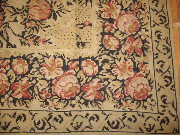 Beige rug corner with pink and red floral motifs and dark blocky outlines resting on wood flooring.