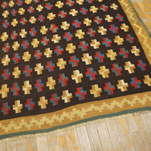 Flat-woven rug with repeating plus signs in color blocks on a dark field and a serrated gold border on worn wood floors.