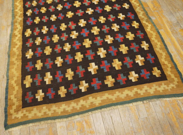 Flat-woven rug with repeating plus signs in color blocks on a dark field and a serrated gold border on worn wood floors.