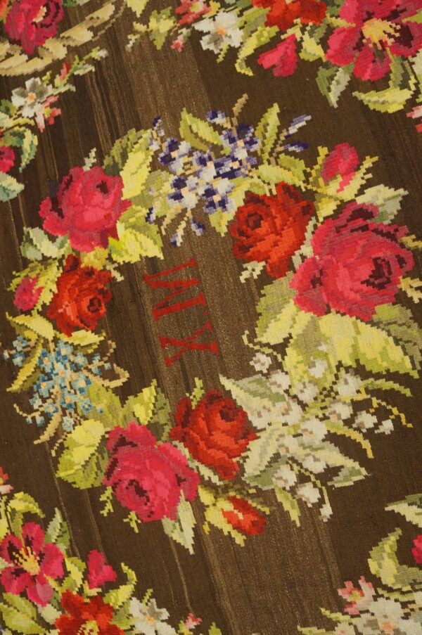 Brown flatweave rug with bright floral wreath design and visible red stitched roman numerals in the center.