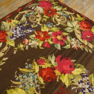 Dark brown low-pile rug featuring a colorful red, purple, and green floral design on worn wood flooring.