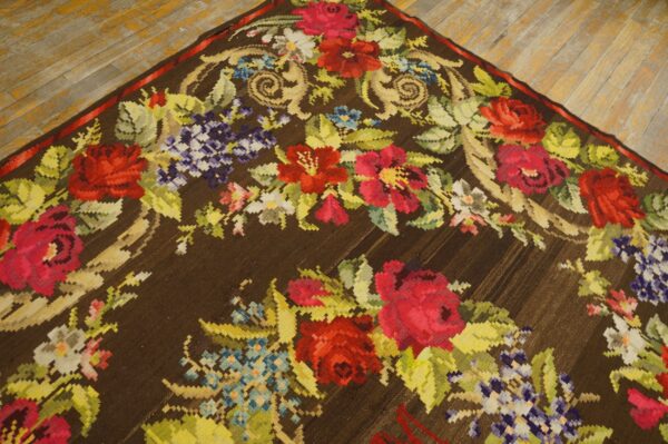 Dark brown low-pile rug featuring a colorful red, purple, and green floral design on worn wood flooring.