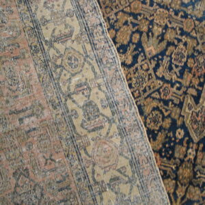 Two antique-style rugs overlap on a hardwood floor, one low-pile navy and gold, the other low-pile pink and beige.