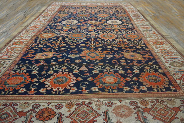 Navy area rug with orange floral motifs and a wide cream border sits on light wood planks.