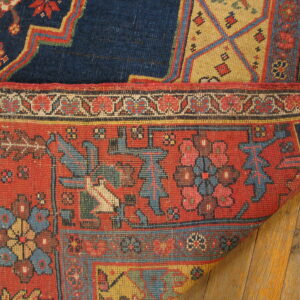 Close up of a red geometric rug folded over a blue rug on warm, wide plank wood flooring.