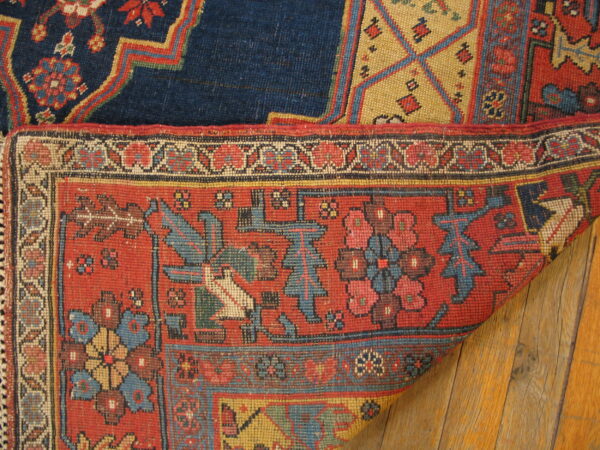 Close up of a red geometric rug folded over a blue rug on warm, wide plank wood flooring.