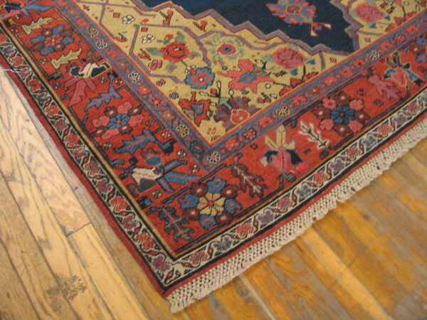 Low-pile rug corner featuring navy, gold, and terracotta colors with intricate borders on light wood floorboards.
