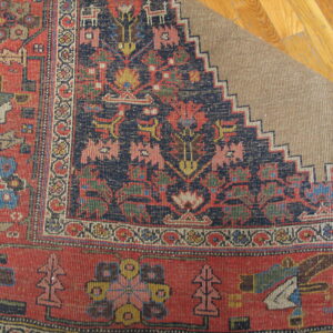 Rust and navy patterned rug corner folded back, revealing a beige backing on light brown hardwood flooring.