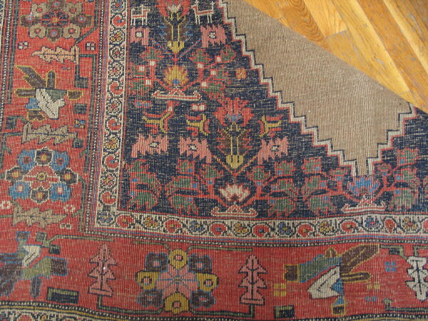 Rust and navy patterned rug corner folded back, revealing a beige backing on light brown hardwood flooring.