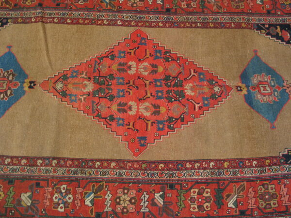 Tan rug center featuring a large, red, stepped diamond medallion bordered by intricate geometric designs and blue accents.