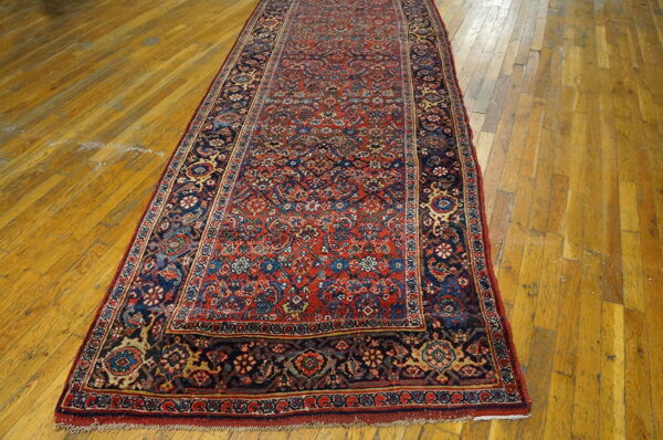 Red and navy traditional runner rug with dense patterns lies on distressed golden hardwood floor.