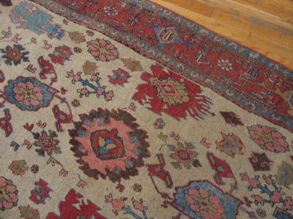 Close-up of a rug with a beige field, red border, and stylized blue, pink, and red floral motifs on wood floor.
