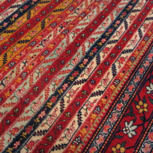 Close detail of a striped rug with red, navy, gold, and pale floral bands next to a red border.