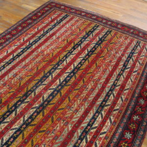 Low-pile rug featuring red, blue, gold, and cream patterned diagonal bands on a wooden floor.