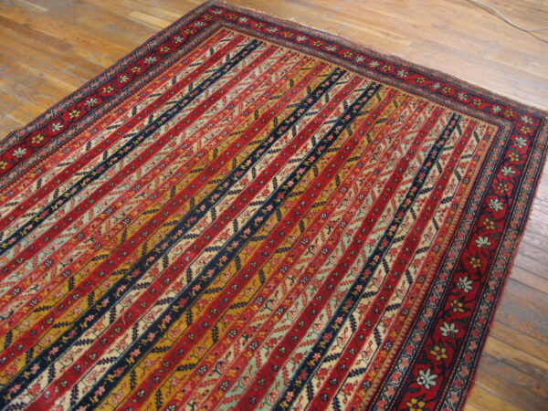 Low-pile rug featuring red, blue, gold, and cream patterned diagonal bands on a wooden floor.