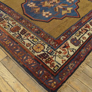 Tan rug corner with complex red, blue, and cream geometric borders on light wood plank flooring.