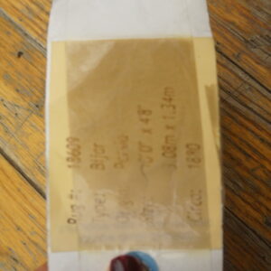 A hand holds a white tag with a translucent tan label against textured brown wood plank flooring.