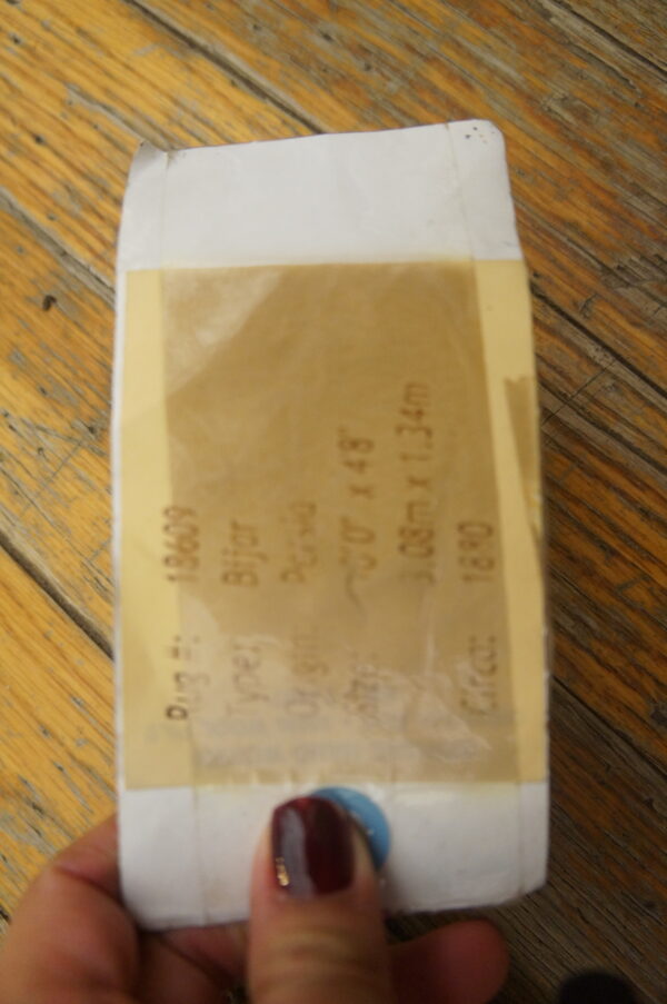 A hand holds a white tag with a translucent tan label against textured brown wood plank flooring.