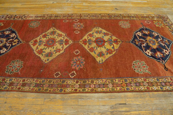 Rust-colored rug featuring repeating yellow and blue patterned diamonds lies on light wooden flooring.