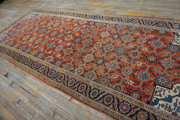 Oversized burnt orange rug with dense floral patterns, wide navy borders, displayed on distressed wooden floor planks.