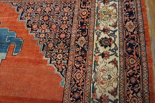 Close-up of a rug featuring an orange field, a stepped blue design, and detailed cream and navy floral borders.