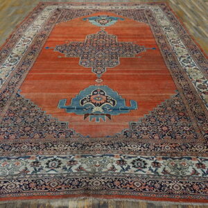 Red-orange rug featuring a large navy blue medallion and complex borders on wood plank flooring.