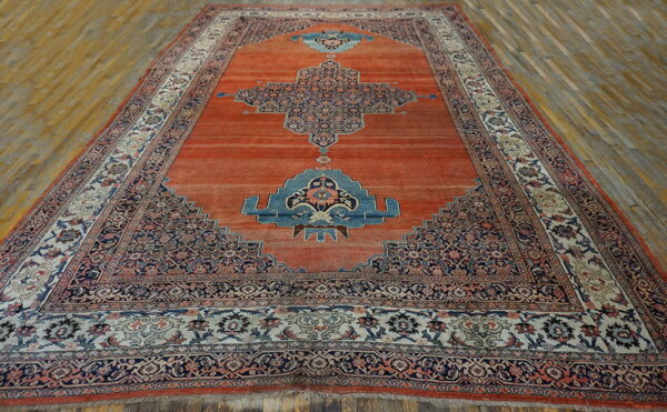 Red-orange rug featuring a large navy blue medallion and complex borders on wood plank flooring.