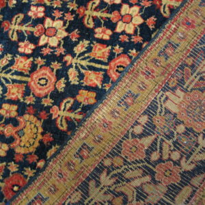 Diagonal view of a navy blue, red, and gold floral pile rug resting next to its woven backing.