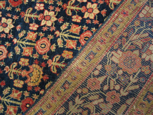 Diagonal view of a navy blue, red, and gold floral pile rug resting next to its woven backing.