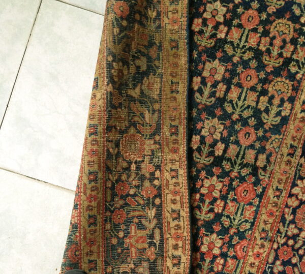 A deep indigo floral rug with red and tan motifs is partially rolled on white tile.