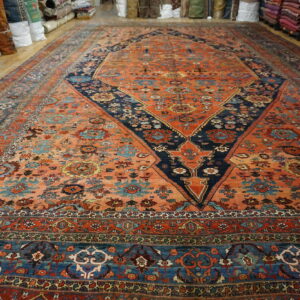 Vibrant orange and blue patterned area rug lying flat on wood flooring, surrounded by stacks of rolled rugs.